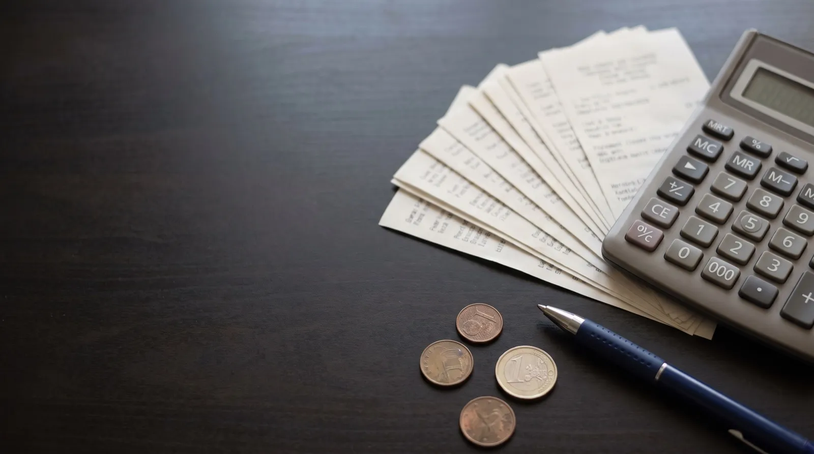 Vista cenital de una calculadora, comprobantes de nómina, monedas y un esfero sobre un escritorio.